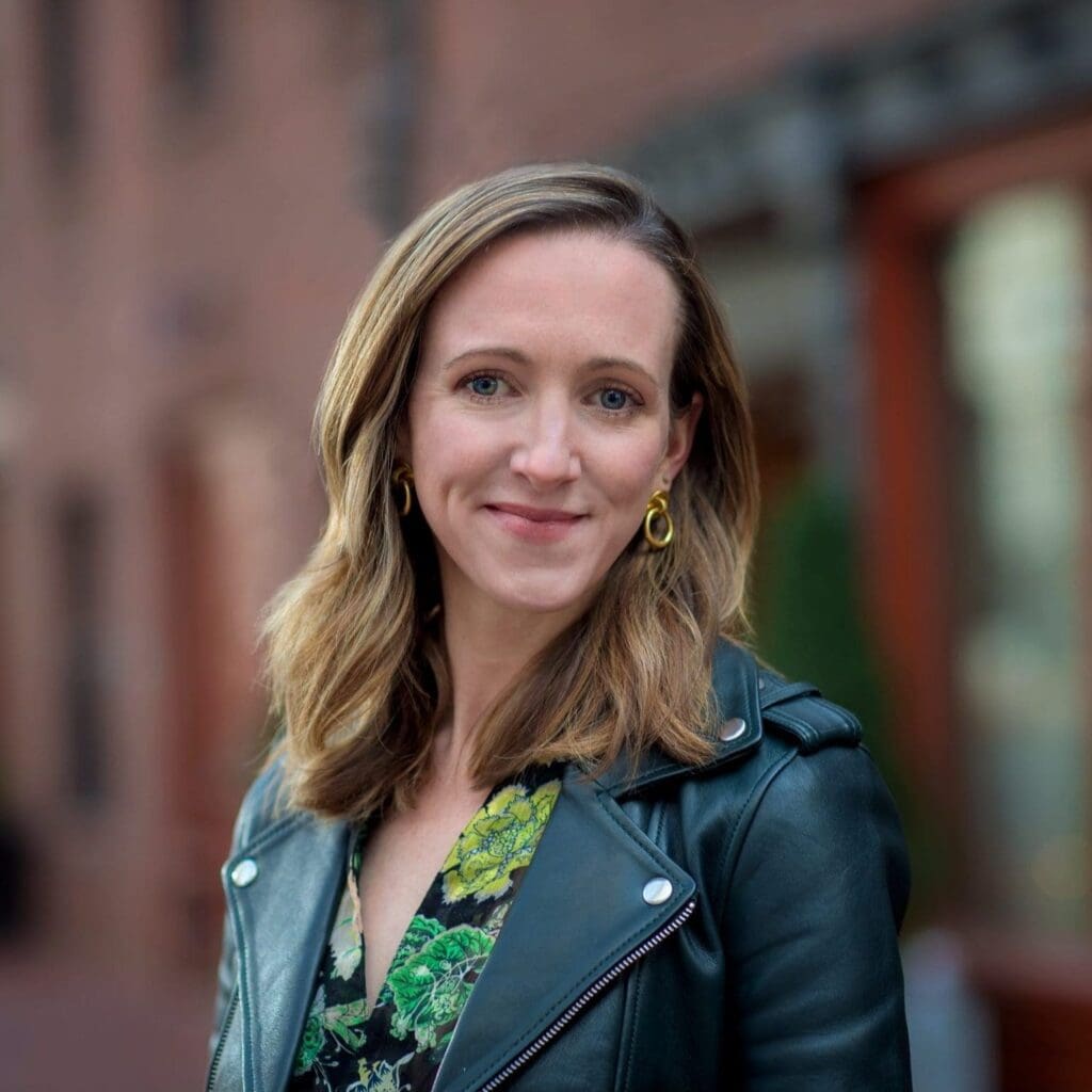 Woman with shoulder-length hair wearing a leather jacket and floral top, standing outdoors with blurred brick buildings in the background.