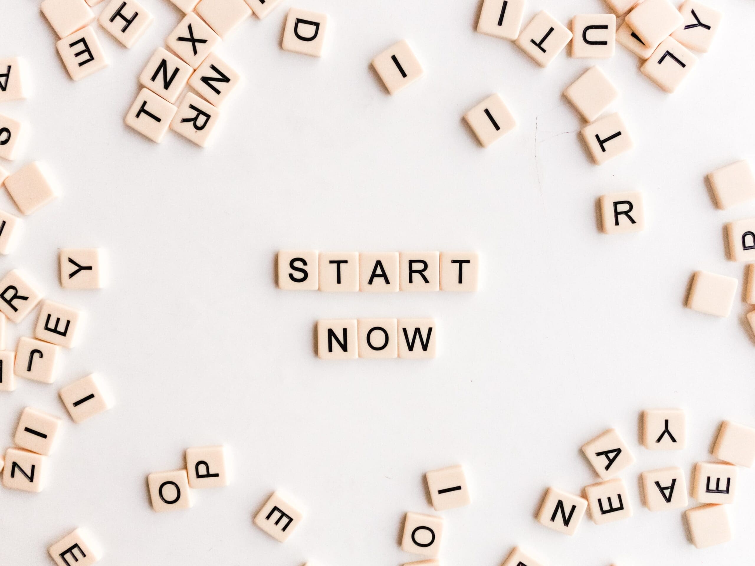 Scattered Scrabble tiles form the words "START NOW" in the center on a white background.